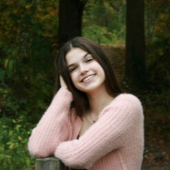 Cailyn smiling while wearing a very light pink sweater with two trees in the background