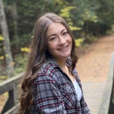 Julia smiling and wearing a plaid red and light blue shirt outside near the woods