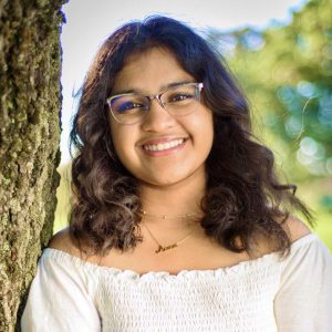 Aanvi smiling and wearing a white shirt next to the side of a tree